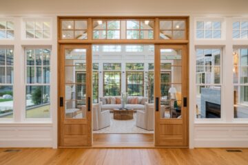 The image depicts a cozy living room in a modern home featuring large windows that flood the space with natural light, a slightly ajar wooden door leading to a lush garden.